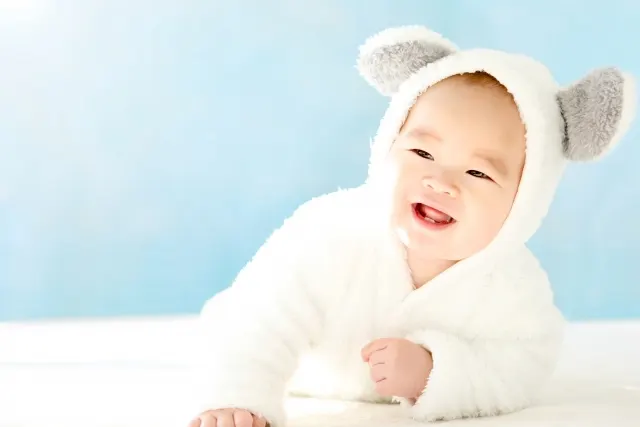 白いふわふわの服を着て、腹ばいで顔を上げて笑う赤ちゃん