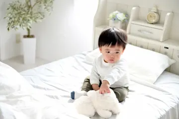 白いベッドの上に座り、ぬいぐるみを手に一人で遊ぶ小さな子ども